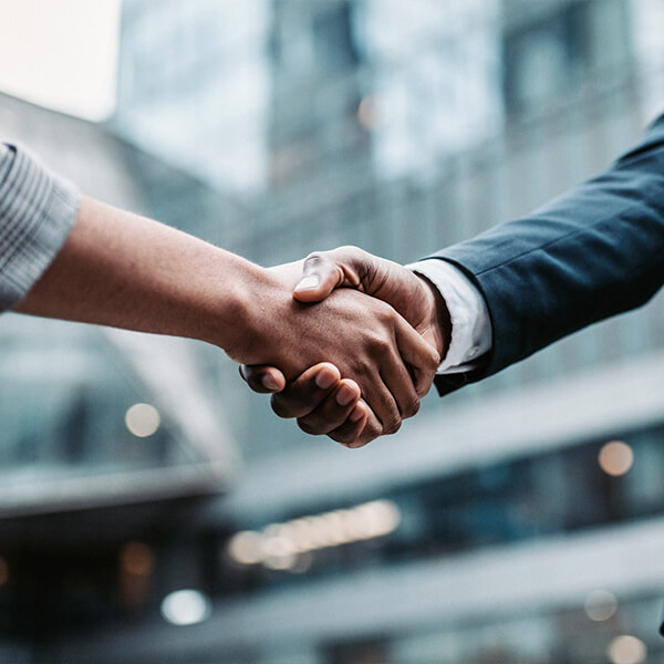 Two people shake hands in front of a blurry office building.